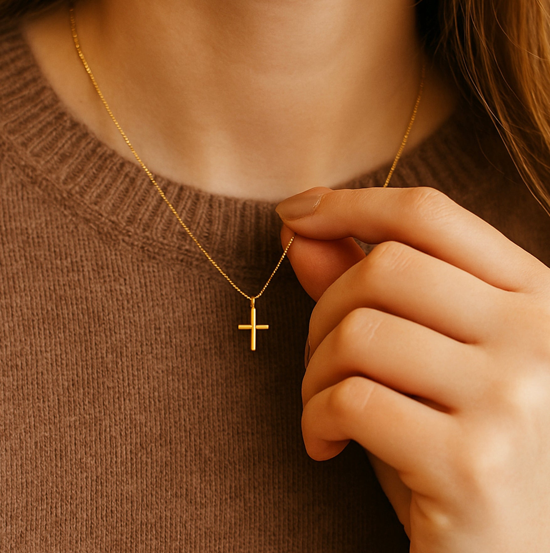 Close-up of woman’s hand gently holding gold cross necklace pendant over brown sweater